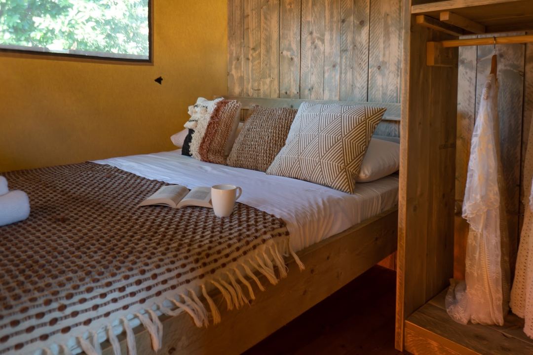 A cozy bedroom with a wooden bed frame, patterned pillows and blanket, featuring warm ochre walls and rustic wooden ceiling details.