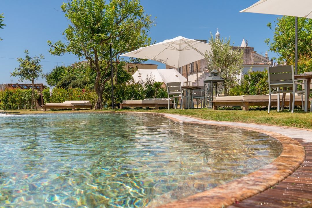 A crystal-clear swimming pool with wooden decking surrounded by white umbrellas, lounge chairs, and lush greenery, with historic buildings visible in the background.
