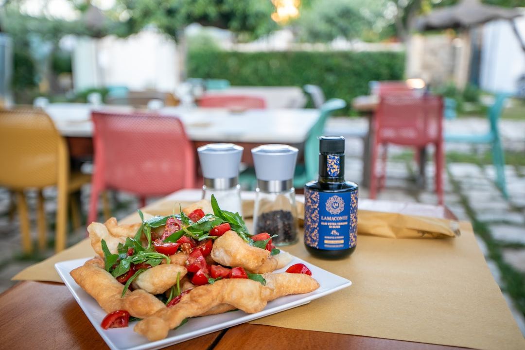 A plate of fried calamari topped with fresh red peppers and arugula, served at an outdoor dining area with colorful chairs and a bottle of wine in the background.