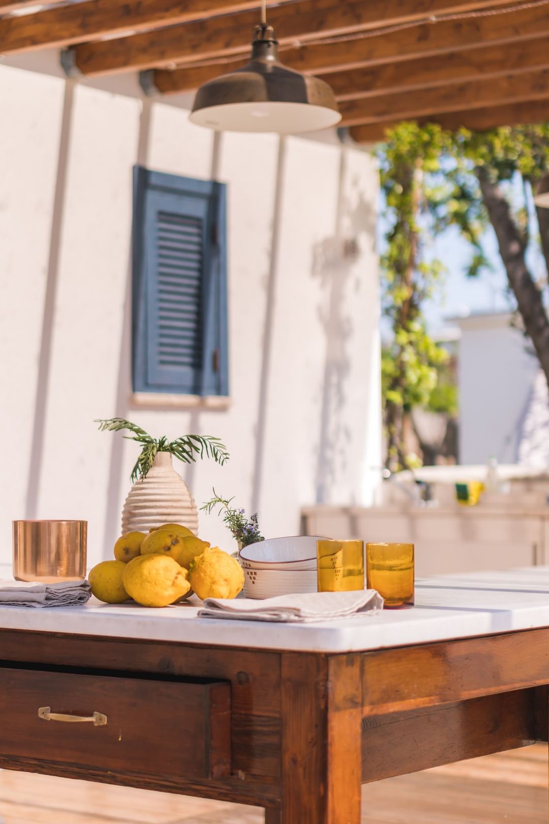 A styled outdoor table setting with fresh lemons, pottery, and glassware under a pendant light on a covered patio with white-washed walls and wooden beams.