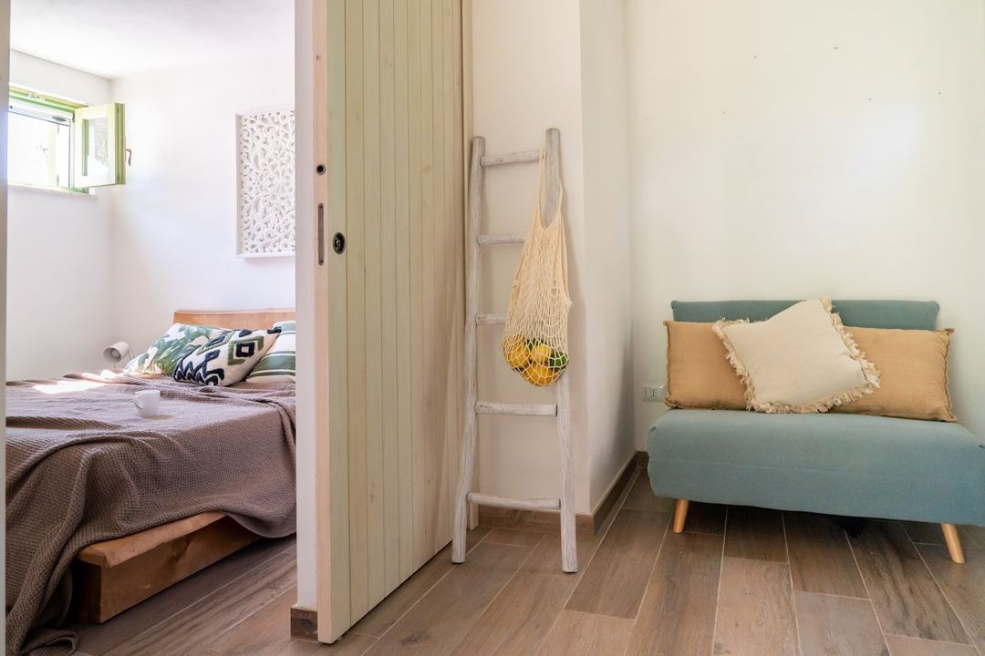 A bright studio room featuring a bed with decorative pillows, a teal sofa with cushions, a wooden ladder with hanging mesh bag, and natural wood flooring.
